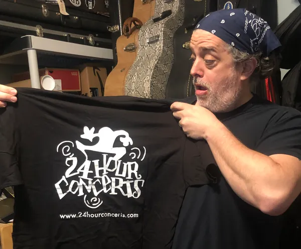 A man holding up a black shirt with the words " 2 4 hour concerts ".